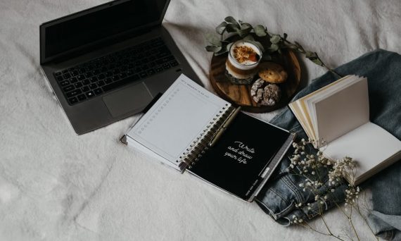A cozy flat lay of a laptop, journals, and snacks on a bed for a relaxed work-from-home setup.